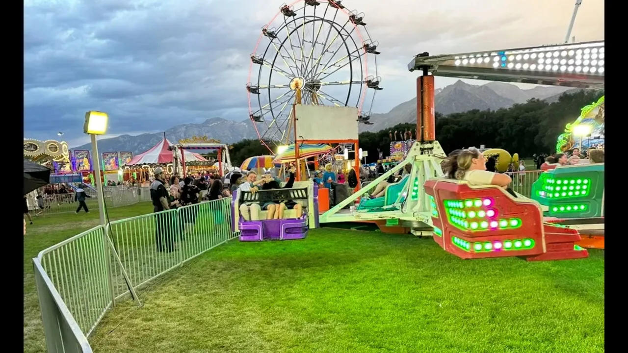 Colorful carnival rides and a Ferris wheel on grass at dusk with crowds and mountains in the background